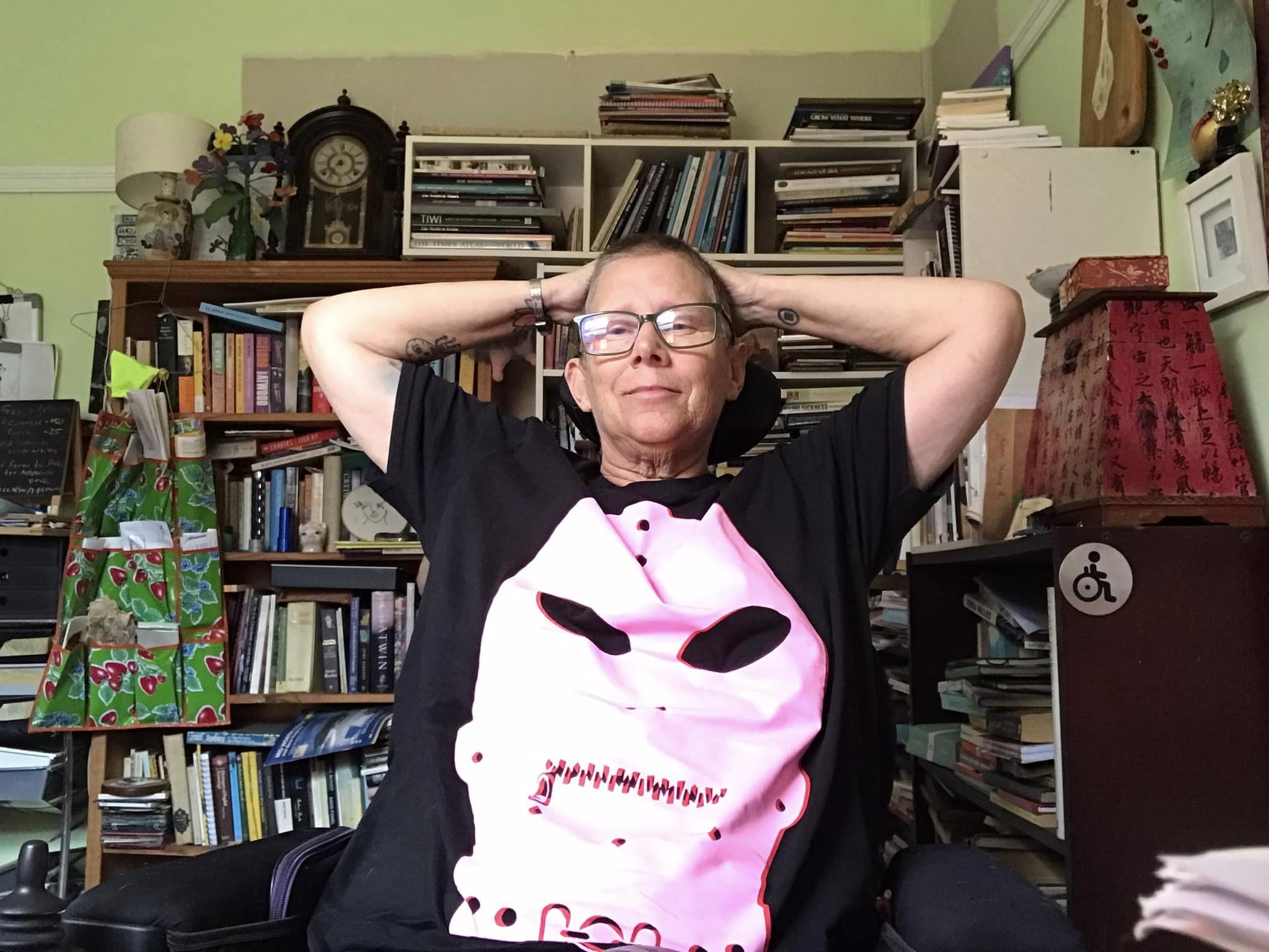 Woman with glasses and a pink gimp shirt leans back with hands behind her head in a room full of books